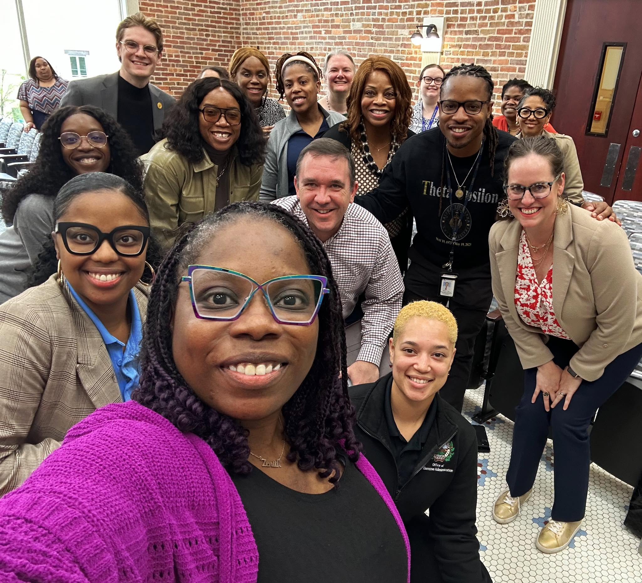 A large group of community partners smiling together for a selfie in a brick-walled meeting room