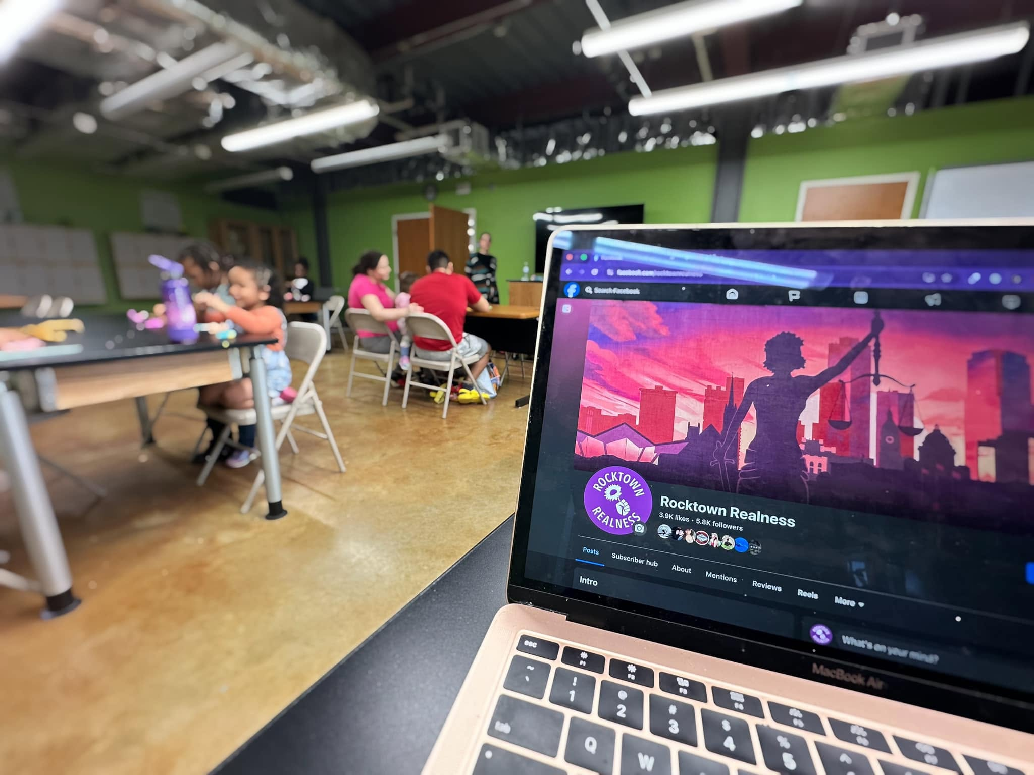 The Rocktown Realness Facebook page open on a laptop in a community room where families gather around tables